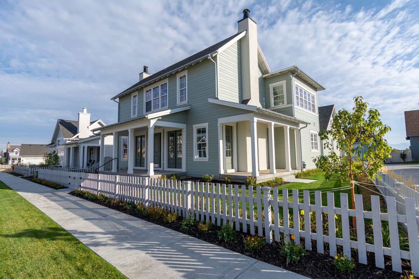 Pale green two-storey house with a deep front porch behind a white picket fence along a sunny sidewalk